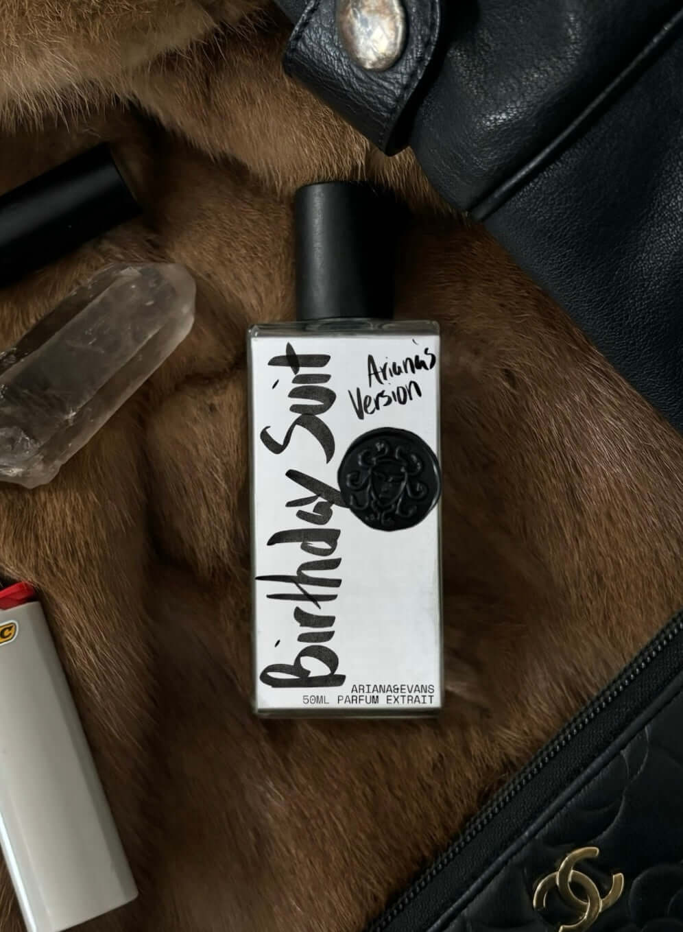 A bottle of 'Birthday Suit' perfume placed on a furry surface, next to a black leather jacket, lighter & spiritual crystal. Indie Niche Fragrances