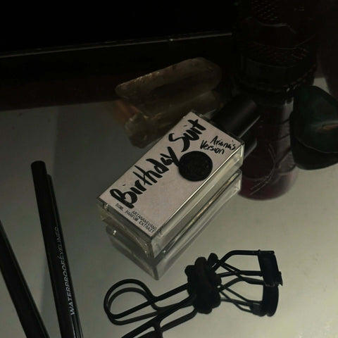 Birthday Suit Parfum in 50 ml glass bottle on a mirror with eyeliner and eyelash curler nearby on side. Ready to party!