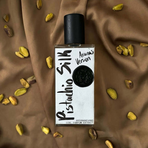 A bottle of 'Pistachio Silk' extrait de parfum by Thalia XOXO, surrounded by scattered pistachios on a brown fabric background.
